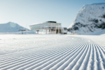 Nahaufnahme einer perfekt präparierten Skipiste mit Gebäuden im Hintergrund.