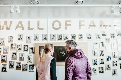 Zwei Personen betrachten die Wall of Fame mit vielen Fotos.