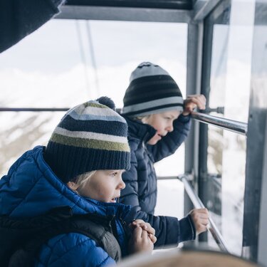 Two children in winter clothing looking at a snowy landscape from a gondola.
