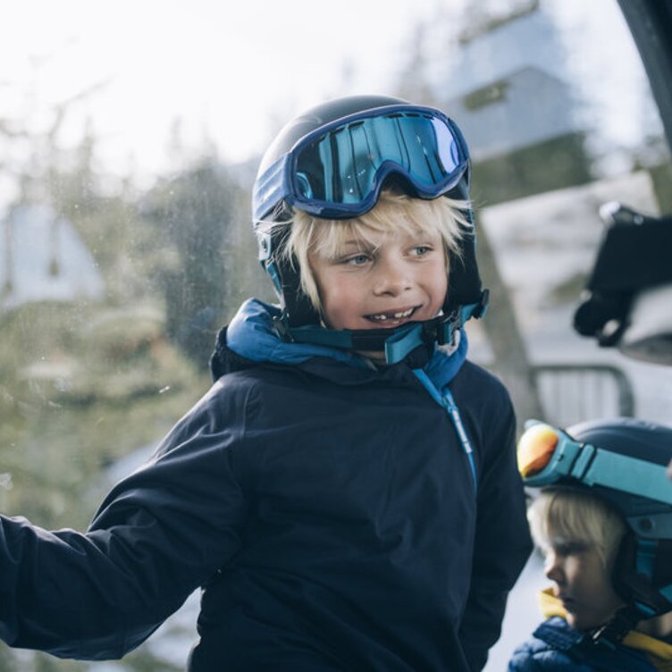 Two children in ski gear smiling inside a gondola during a ski vacation.