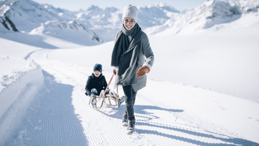 Woman pulling child on a sled through snowy mountain landscape.