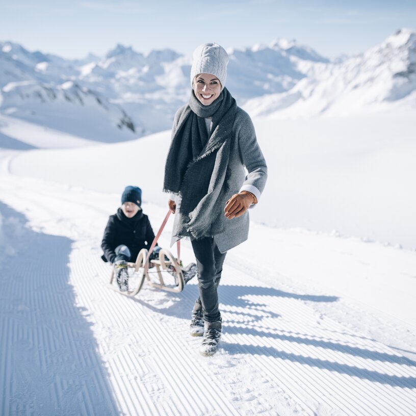 Woman pulling child on a sled through snowy mountain landscape.