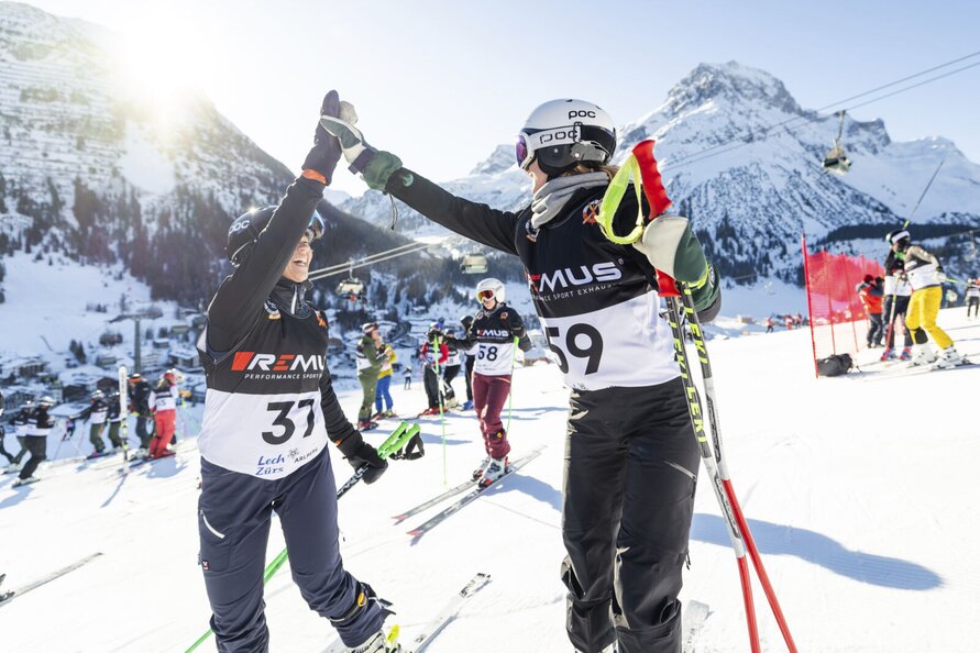 Two skiers high-five each other at a ski race in the mountains, with other skiers in the background.