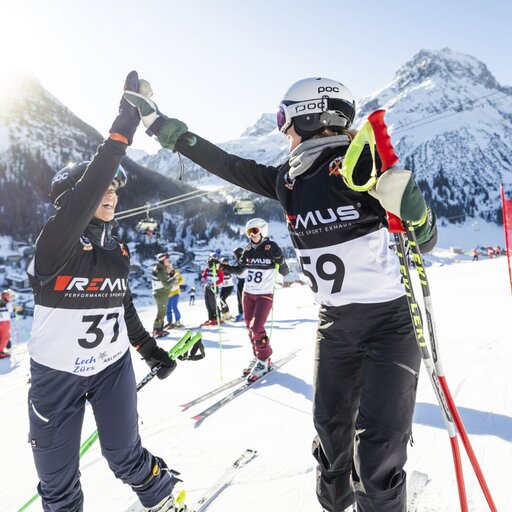 Zwei Skifahrer geben sich ein High-Five bei einem Skirennen in den Bergen, mit weiteren Skifahrern im Hintergrund.