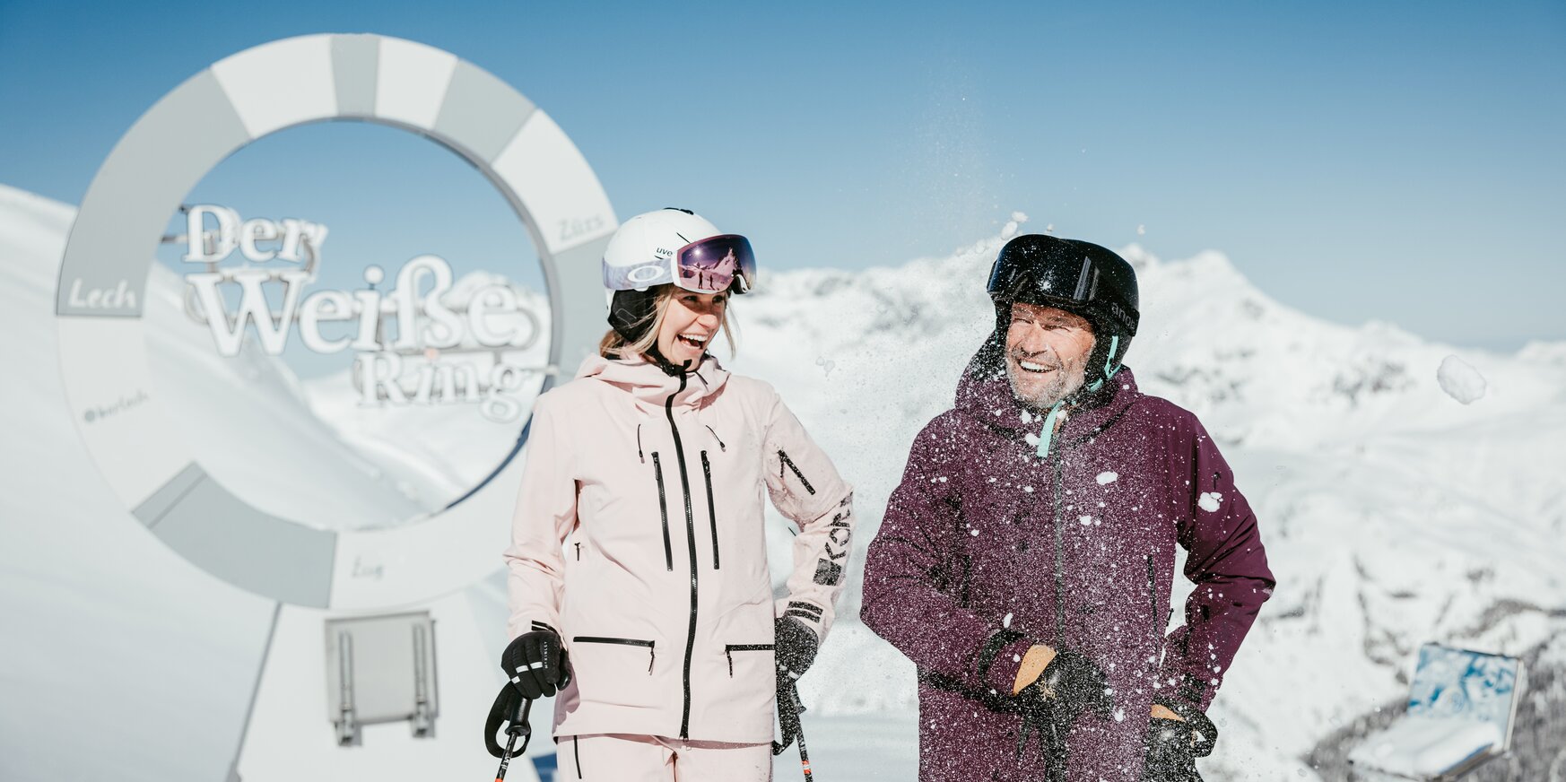 Two skiers laughing in front of the 'Der Weiße Ring' (The White Ring) sign in the snowy mountains.