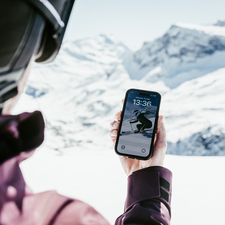 Eine Person mit Skihelm schaut auf ein Smartphone, am Display wird die Uhrzeit 13:36 angezeigt.