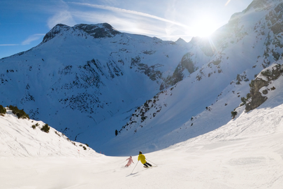 Two people skiing on a sunny mountain slope with snowy peaks in the background.