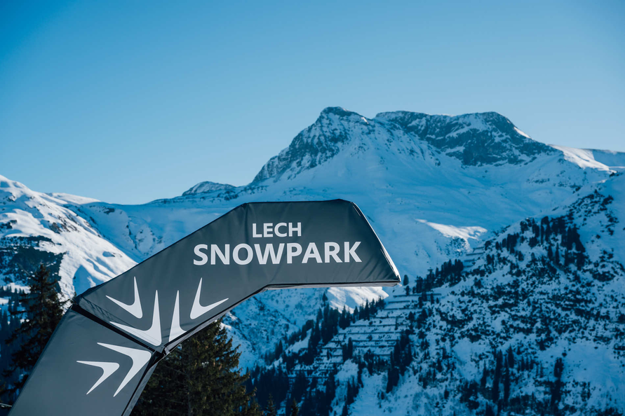 Lech Snowpark Schild vor verschneiten Bergen und blauem Himmel.