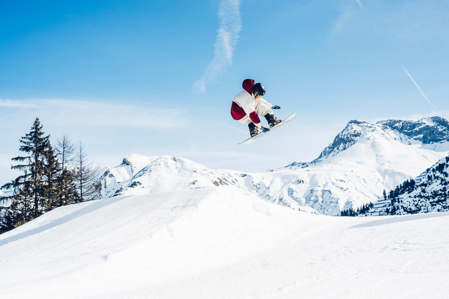 Snowboarder springt über eine Schneerampe mit Bergkulisse.