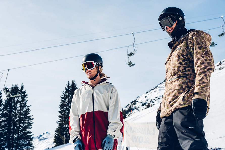 Zwei Männer in Wintersportkleidung, möglicherweise Skifahrer, mit Helm und Skibrille vor schneebedeckter Landschaft und Skilift.