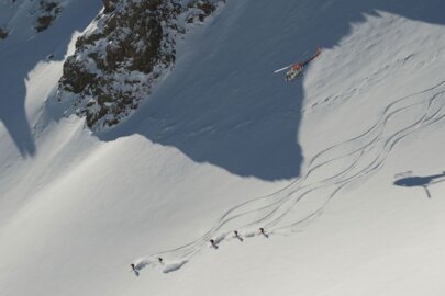 Several skiers are descending a steep slope, accompanied by a helicopter.