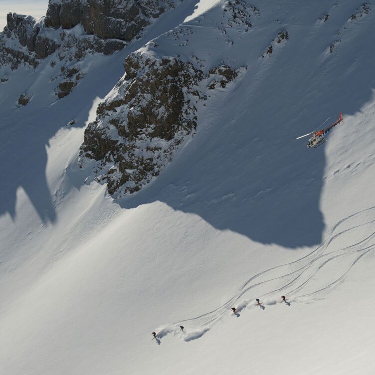 Mehrere Skifahrer fahren einen Steilhang hinunter und werden von einem Hubschrauber begleitet.