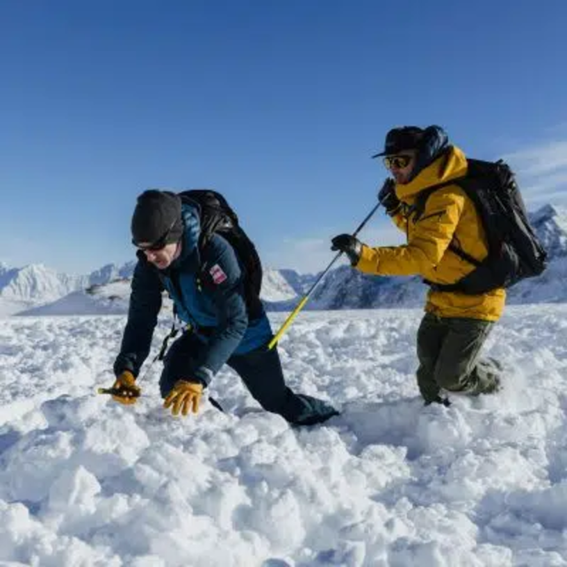 Two people at an avalanche training area with a mountain backdrop.