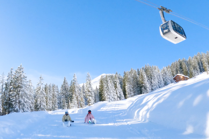 Two people sledging in the snow, with a cable car in the background.