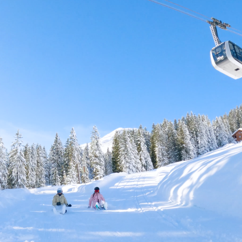 Two people sledging in the snow, with a cable car in the background.