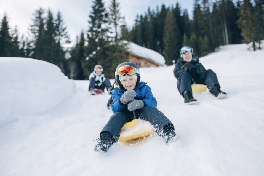 A family sledging down a snowy hill, surrounded by trees.