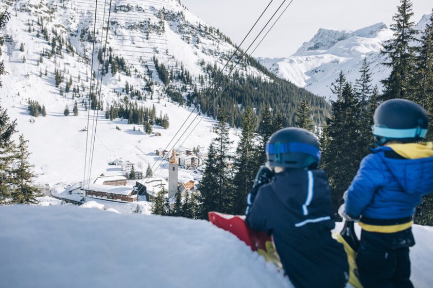 Two kids with helmets sitting in the snow overlooking a snowy village.