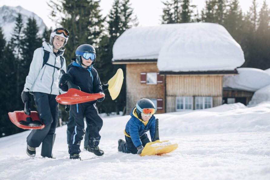 Family sledging in the snow in front of a cabin.