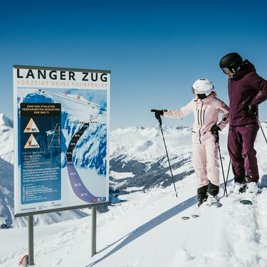 Zwei Skifahrer stehen vor einem Warnschild mit Informationen zur Piste Langer Zug in den Alpen.