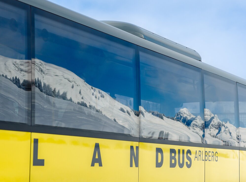 A yellow bus and snow-covered Alps reflected in the windows.