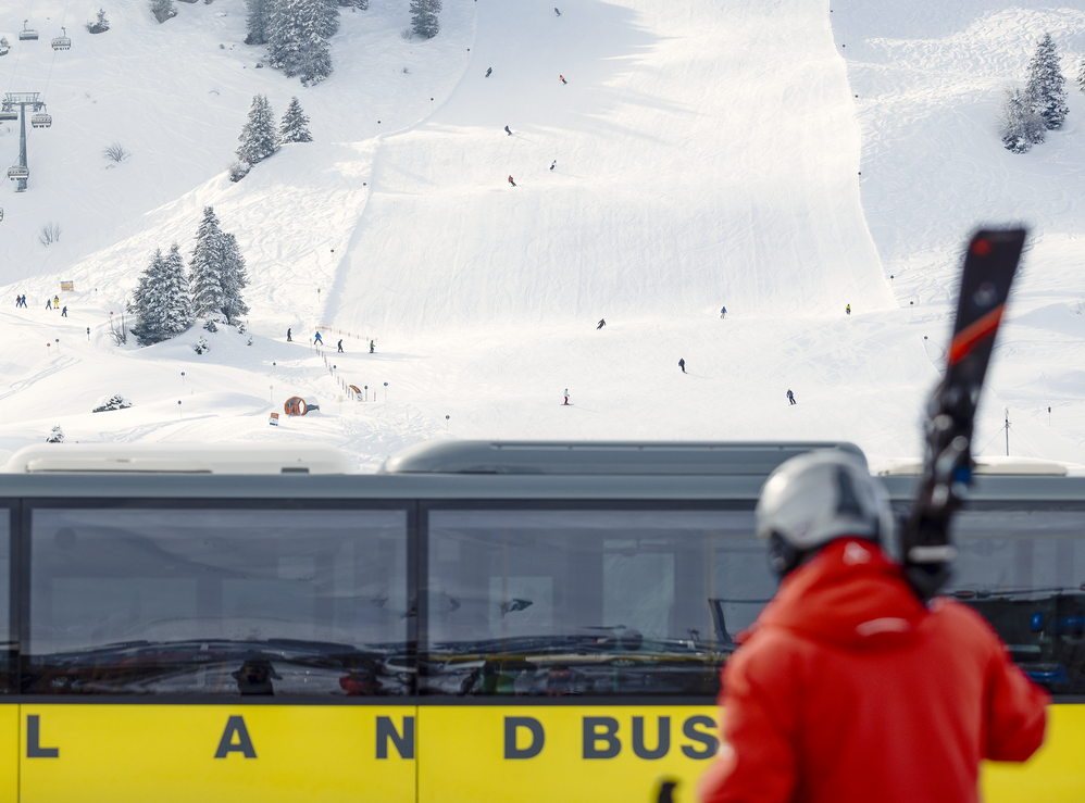 A person in a red jacket with skis in front of a yellow bus. Ski lift and skiers in background.