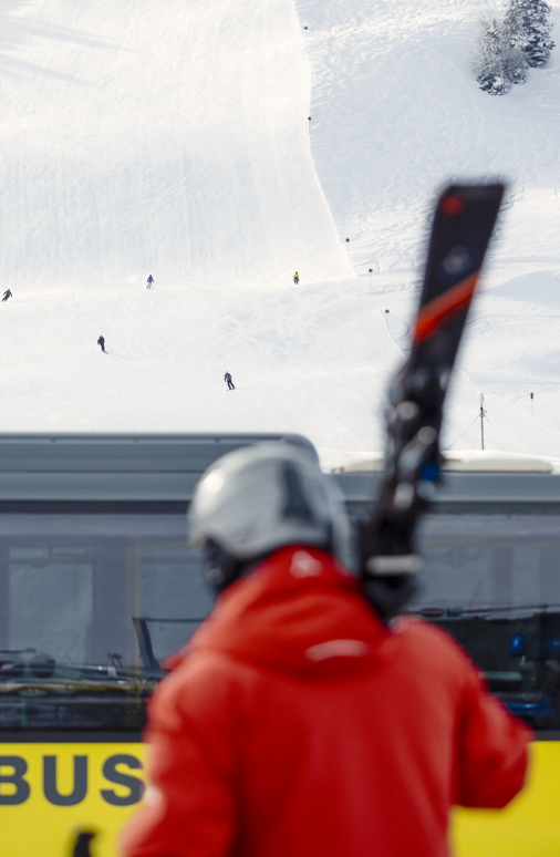 Person in roter Jacke und Skiern über der Schulter vor einem gelben Bus, der neben einer Piste parkt.