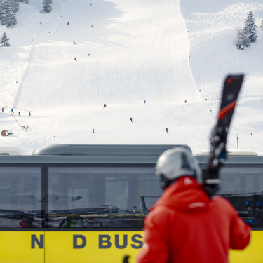 A person in a red jacket with skis in front of a yellow bus. Ski lift and skiers in background.