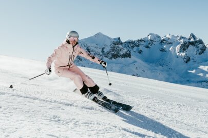 Person in rosa Skianzug fährt elegant einen Hang hinab vor einer verschneiten Bergkulisse.