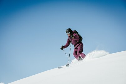 Person skiing down a snowy slope in a purple suit under a clear sky.