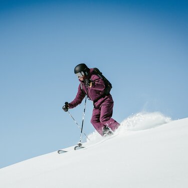 Person skiing down a snowy slope in a purple suit under a clear sky.