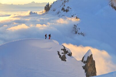 Zwei Personen stehen auf einem schneebedeckten Berggipfel. Unter ihnen hat sich eine Wolkendecke breit gemacht.