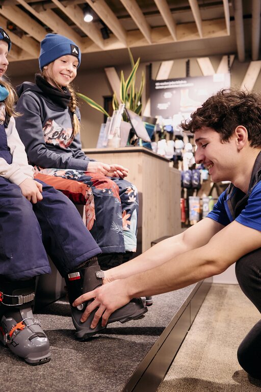 Two children in snow gear trying on ski boots in a shop.