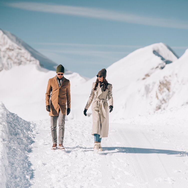 Zwei Personen spazieren auf einem schneebedeckten Bergweg, umgeben von verschneiten Gipfeln.
