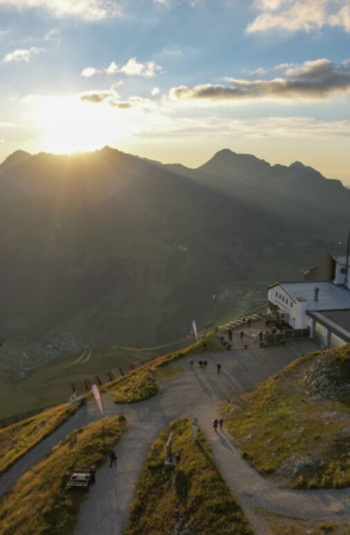 Menschen genießen den Sonnenuntergang auf einem Berg mit Seilbahn und Hütten im Hintergrund.