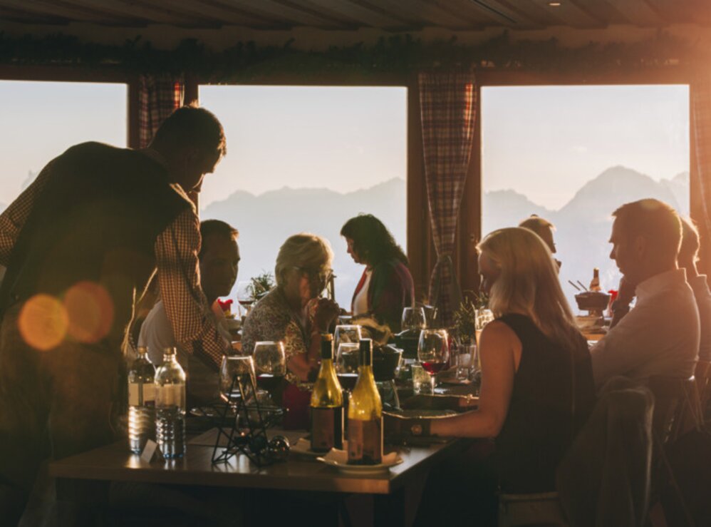 People at a table, drinking wine, enjoying sunset with mountain silhouette in the background.
