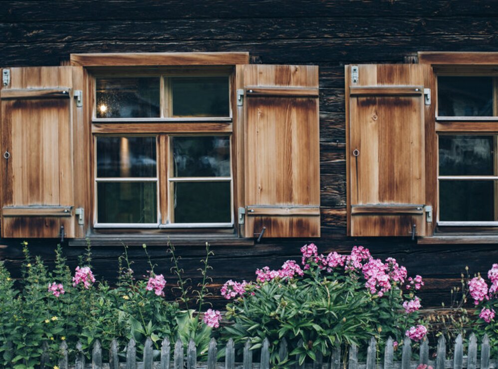 Zwei Holzfenster mit offenen Fensterläden und rosa Blumen davor.