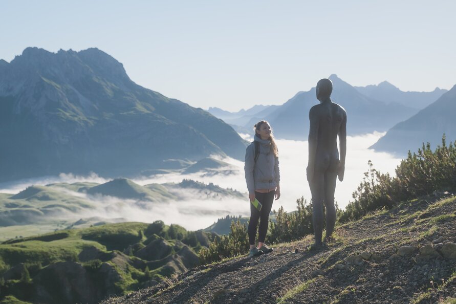 A woman stands beside a sculpture in a mountain landscape.