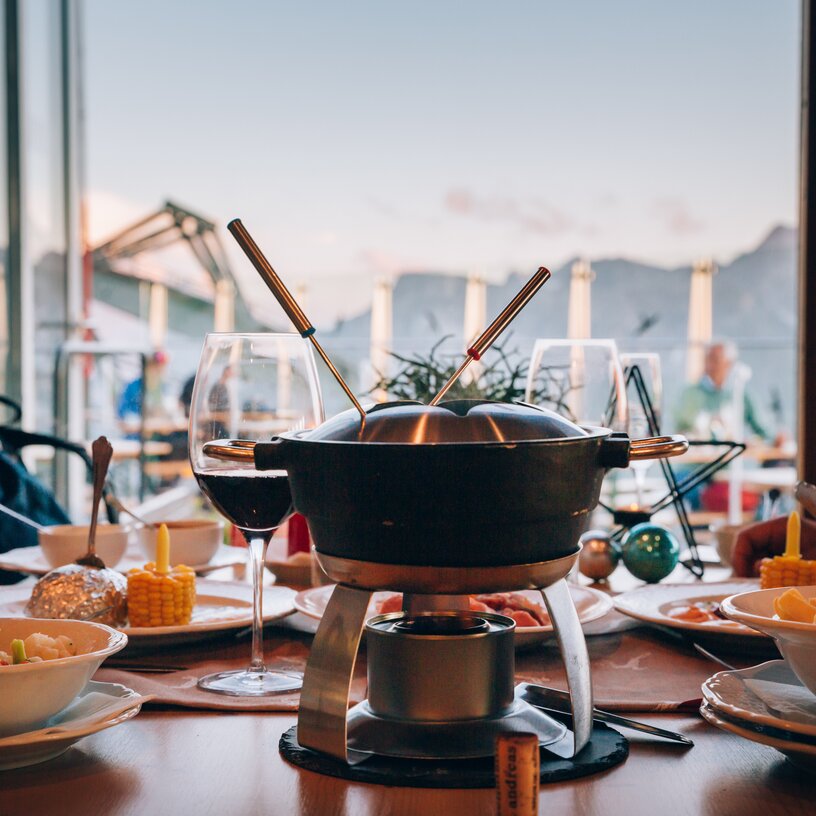A table with cheese fondue, wine, and sides against mountain backdrop.