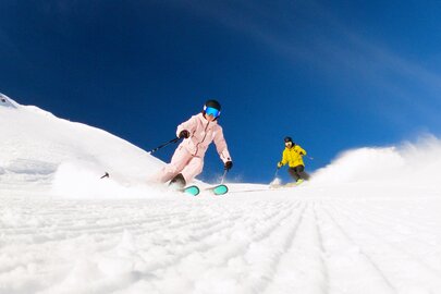 Zwei Skifahrer in bunten Anzügen fahren eine schneebedeckte Piste hinunter.