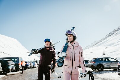 Zwei Skifahrer tragen ihre Skier über einen Parkplatz eines Skigebiets.