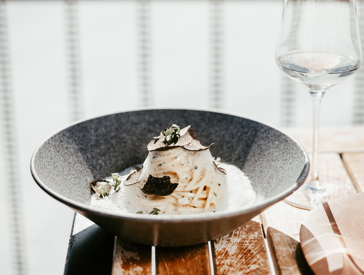 Ein Teller Pasta mit Rahmsauce und Trüffelscheiben, neben einem Glas Weißwein auf einem Holztisch.