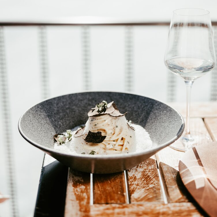 A plate of pasta with cream sauce and sliced truffle, next to an empty wine glass on a wooden table.