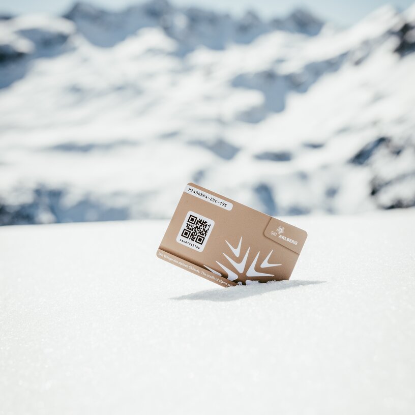 Goldene Gutscheinkarte von Ski Arlberg mit QR-Code im Schnee vor Bergkulisse.