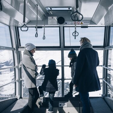Four people in a cable car with a view of snowy mountains.