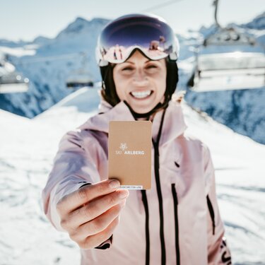 Woman on ski slope holding a Ski Arlberg pass against snowy background.