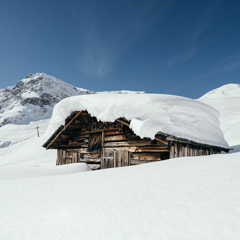 A small wooden cabin is buried in deep snow, set against a snow-covered mountain and blue sky.