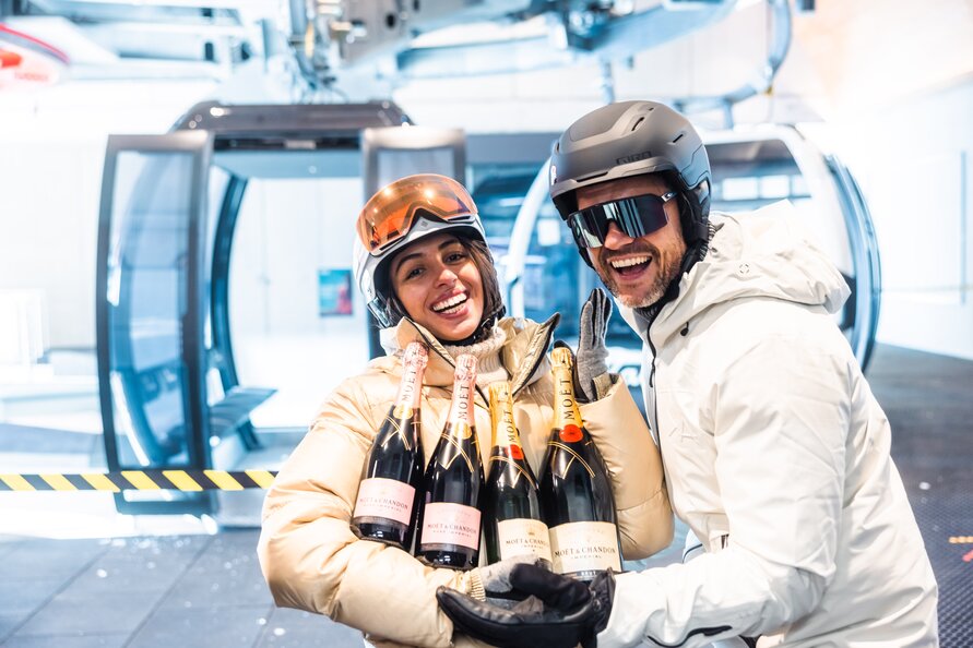 Ein gut gelauntes Pärchen in Skiausrüstung hält 4 Champagnerflaschen vor sich. Im Hintergrund ist eine Gondel zu sehen.