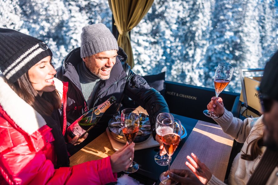 Gruppe fährt in einer Gondel und prostet sich mit Champagner zu. Draußen ist eine verschneite Winterlandschaft zu sehen.