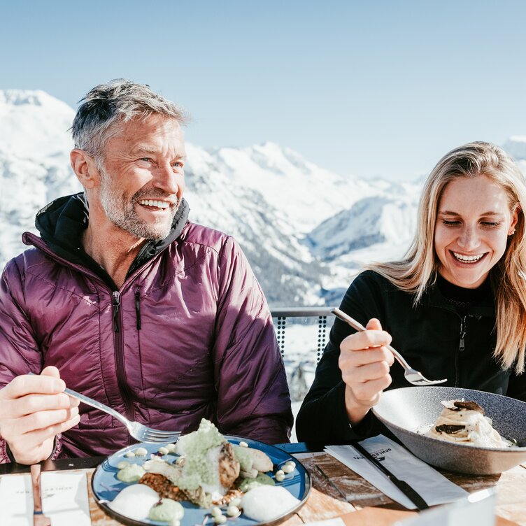 Lächelndes Paar genießt Mittagessen auf einer Skihütte bei herrlichen Wetter und Bergpanorama.