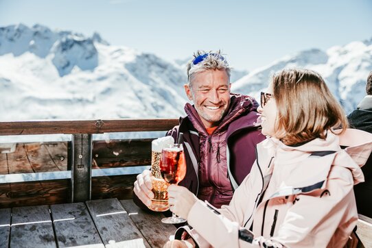Ein Paar genießt Getränke auf einer Terrasse mit schneebedeckten Bergen im Hintergrund.